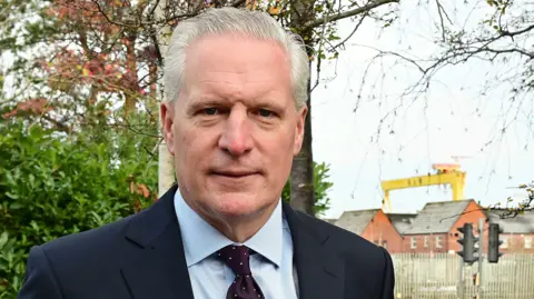Pacemaker Alan Stout. He has short grey hair, wearing a navy blazer, light blue shirt and navy polka dot tie. The Harland and Wolf cranes are in the background.