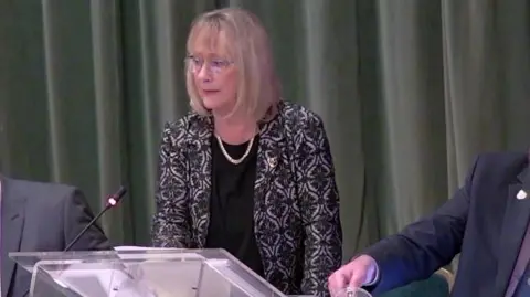 Councillor Brenda Warrington has a blonde hair, a black dress, a necklace and a patterned jacket and is speaking at the lectern in a Tameside Council meeting