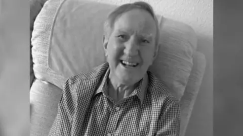 Geoff Peters Neville Foulger smiling as he sits in an armchair in a checked shirt