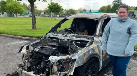 A woman in a blue fleece and leggings stands next to a burnt-out car. There is some green space behind her. 