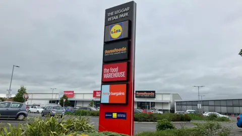 A colourful vertical sign listing shops on a retail park including Lidl, Farmfoods, the food warehouse and home bargains. The sign is on the edge of a car park and the shops are in the background on an overcast day.