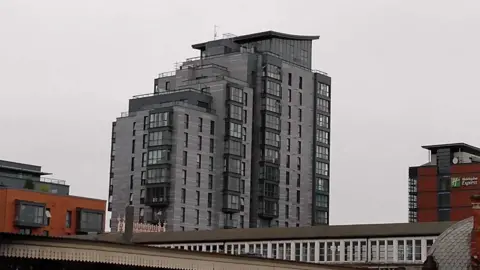 A modern-looking grey and black tower block, significantly taller than any of the other buildings around it, against a white cloudy sky.