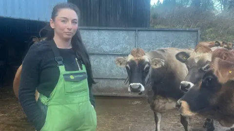 A woman wearing farm overalls looking solemn whilst standing next to her herd of cows.