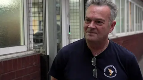Tony Miller is wearing a navy t-shirt, with the Whitechapel Mission logo on it. He is standing outside the Mission, on the street
