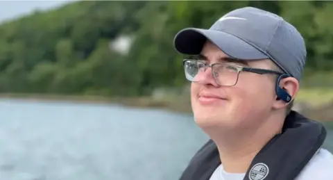 A smiling man wearing a cap and spectacles is pictured in close up and in profile. He is outdoors, and behind him is a large body of water, with a tree-lined shore. He also wears music earphones and has an inflatable life preserve around his neck and shoulders