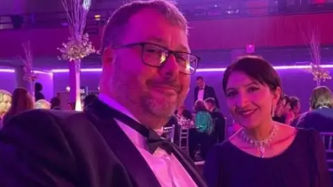 University of Bradford A close-up of a man with a beard, wearing glasses and a suit with a bow tie. He is pictured alongside a smiling woman wearing a necklace and dress