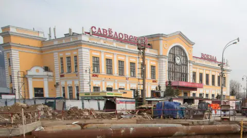 A yellow and white building with red lettering in Russian stands above construction works.