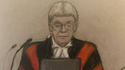 Julia Quenzler Judge Mark Lucraft, who is wearing red and black judges robes, a white legal wig and glasses. He is looking at a tablet in front of him and there are two microphones to the side