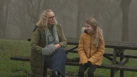 BBC/Luke Manterfield A picture of Cheryl Blenkinsop (left), with blonde hair, glasses and a green coat, sitting on a picnic bench next to her daughter Eliza (right), who has long blonde hair and is wearing an orange coat. There are trees in the background.
