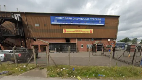Google A large building with red bricks and brown metal exterior. There is a metal fence surrounding a small car park in front of it. A large blue sign on the building reads "Perry Barr greyhound stadium". A sign below it reads "the home of the Birmingham Brummies speedway team".