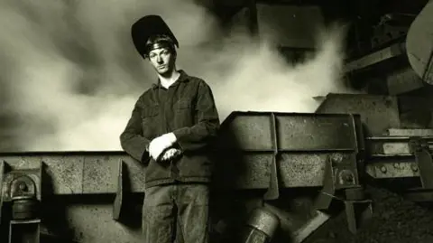 Steve Orino A black and white photograph of a foundry worker leaning on a piece of machinery. The man looks quite young and is wearing a safety visor on his head which is tipped up, revealing his face. The background appears to be smoky.