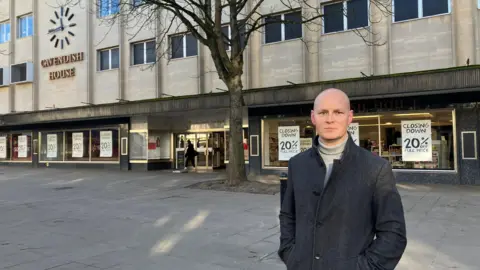 A bald middle aged man is wearing a long dark grey coat with a pale turtleneck beneath it as he looks sternly at the camera outside a department shop called Cavendish House on a winter's day. 