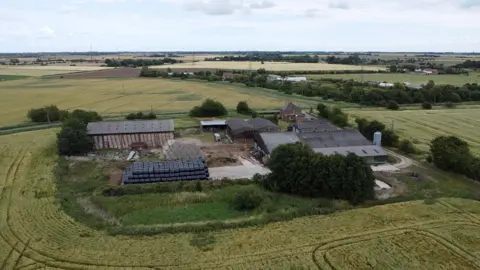 The Homer's farm seen from drone footage. There is a farmhouse surrounded by an assortment of farm buildings and several bales of hay. The plot is surrounded by fields and open countryside.