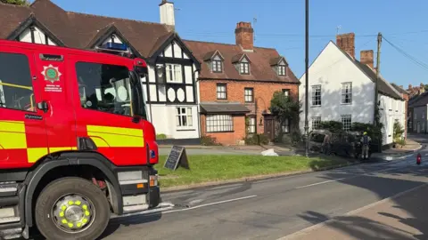 Uttoxeter Fire Station A fire engine is positioned on the left hand side of the image, with the burned carcass of a campervan in a shaded area to the right hand side of the image. There are firefighters attending to the vehicle. There is a white building visible nearby, along with a brick building and a black and white building alongside it.