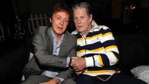 Getty Images Sir Paul McCartney and Brian Wilson shake hands. They are both seated in a dark room and looking at the camera. Sir Paul is wearing a grey suit and blue striped shirt and Wilson has a black, yellow and white long-sleeved polo shirt.
