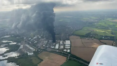 David Gill An aerial view of the fire, taken from an airplane, showing a towering plume of black smoke rising into the clouds above. Fields and the nearby town of Rushden are visible surrounding the epicentre of the fire.