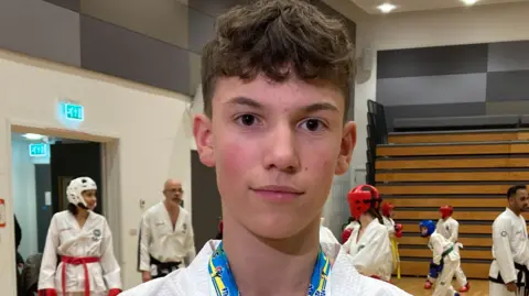 A teenage boy stands in a martial arts training hall. Medal ribbons can be seen round his neck. Other martial artists are training behind him.