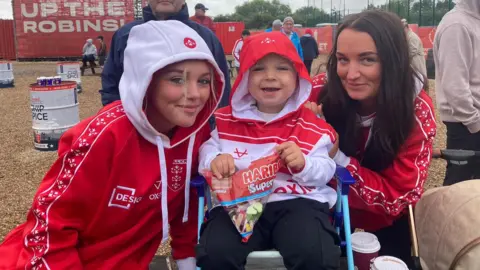 BBC/Sally Fairfax Three Hull KR fans wearing red and white hooded sweatshirts. The woman on the right has long brown hair and is holding on to the back of a deck chair on which a child, who has their hood up, is sitting. The child is in the middle. On the left is a young girl who also has a hood up over her head. All three are smiling at the camera.