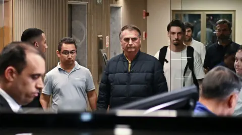 ANDRE BORGES/EPA/Shutterstock Former Brazilian President Jair Bolsonaro is wearing a navy blue parka as he leaves a building with several people around him. He is seen through glass doors leaving a lobby with elevators behind him. He has a grim expression.