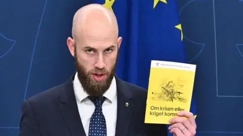 Getty Images Swedish Defense Minister Carl-Oskar Bohlin holds a copy of the new version of the preparedness manual "When disaster or war comes". This pamphlet has a yellow cover and a picture of a tired woman in the army holding a big gun.