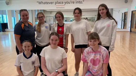 Seven girls aged between 11-14 stand smiling in a group with their instructor in an exercise studio. They are wearing sport kit. 