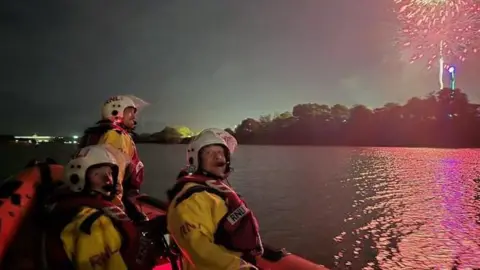 Three RNLI personnel off the Isle of Wight coast at night in a RIB, with fireworks going off behind
