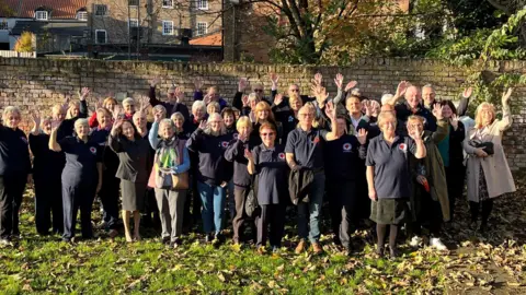 BBC/ Julia Lewis A group of men and women stood on grass, in front of a brick wall. They use one hand to wave towards the camera.