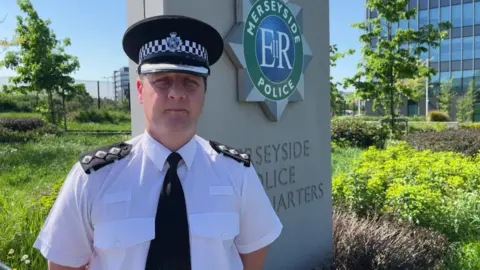BBC/Jonny Humphries Ray Mavrakakis, in his police uniform of a white shirt, black tie and black police cap, stands next to a stone monument carrying the crest of Merseyside Police and the words Police Headquarters