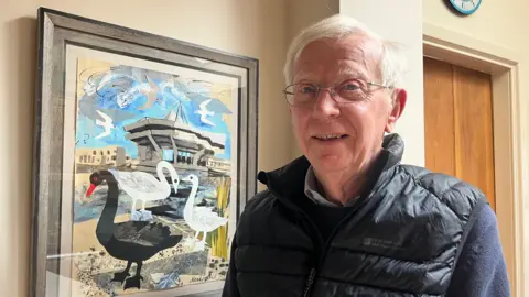 A man with short white hair smiles as he stands in light-filled room in front of a playful painting of sea birds in a seaside location. He is wearing glasses, a black gillet and a navy-blue fleece.