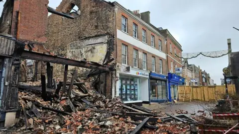 Fire-gutted building with debris spilling out onto a street of terraced buildings a wooden fence is across the street to the right.