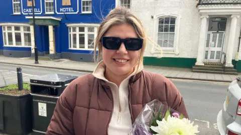 Kayla Lovell pictured in the street wearing sunglasses and a brown puffer jacket and cream sweathshirt. She has blonde hair in a pony tail and is holding a bouquet of flowers. The Castell hotel is behind her with a blue facade