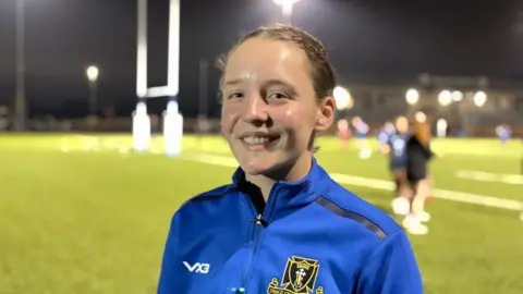 Anya Steel smiles at the camera with her brunette hair pulled back into a ponytail. She's wearing a light blue jacket with the Dings Crusaders RFC logo on the right side. Behind her is a blurred image of a rugby game.