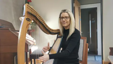 Clara Wilson is sitting in a church with a large harp. Her two hands are playing the strings. She's looking into the camera. She wears glasses and her hair is blond. She has black clothes on. An organ is beside her. 