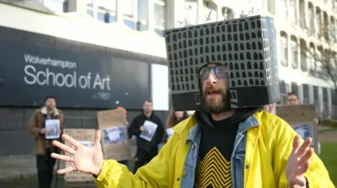 A man wears a bright yellow coat over a denim jacket and patterned t-shirt. On his head, he wears a cardboard box painted grey and black - a replica of the School of Art building. He gestures with his hands while making a comment