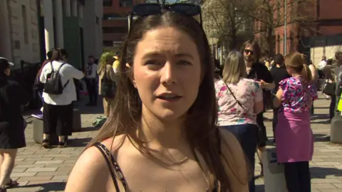 Eva Martin is looking into the camera. She has long brown hair. Sunglasses are on top of her head. People are standing in the background behind her, 