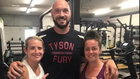 Jade, a blonde woman and another woman with brown hair stand in a gym on either side of Tyson Fury, who wears a Tyson Fury t-shirt, he is bald and has a beard.