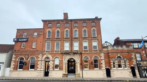 A wide-angle view of a large red-brick building with arched doorways and tall windows. The building has decorative stone detailing and multiple floors, with neighbouring shops on either side. A sign on the top left of the building depicts the University of Lincoln crest and the word "Barbican" in white text set against a black background. The sky above the building is overcast and cloudy.