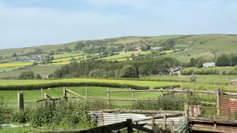 Rolling green hills stretch across the horizon under a pale blue sky. Patchwork fields divided by hedgerows and clusters of trees fill the middle distance, with scattered farm buildings dotted along the slopes. A wooden post-and-rail fence and corrugated metal sheeting line the front of the scene.