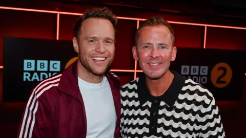 Olly Murs is standing on the left wearing a white T-shirt and red jacket with stripes down the arms. He has short brown hair and is smiling at the camera. On the right is Scott Mills on the right wearing a black and white zigzag T-shirt and short brown hair, and is smiling towards the camera. In the back is a sign that says BBC Radio 2.  