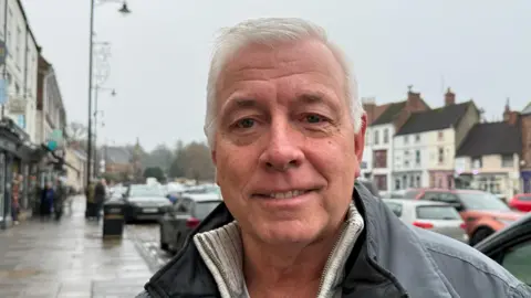 Geoff Swainston is standing next to a car on a busy street. He has short white hair and is wearing a thick grey coat over a grey sweater.