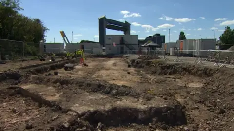 The site of an archaeological dig with a large area of uncovered earth and the outline of low walls under a blue sky. In the background are portable buildings and a large tidal barrier.