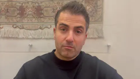 A man with short dark hair in a black top looks at the camera