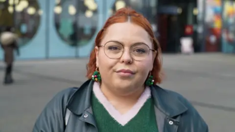 Jamie Niblock/BBC Sophie Kirkham has died red hair, glasses, and is wearing a green knitted jumper and a leather jacket. She is standing outside looking at the camera.
