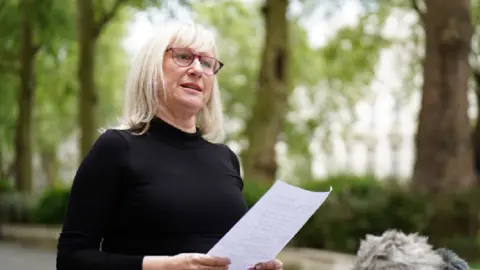 Anna-Louise Marsh-Rees, from the Covid-19 Bereaved Families for Justice Cymru campaign, speaks to the media outside to the UK Covid-19 Inquiry at Dorland House in London, during its first investigation examining if the pandemic was properly planned for and "whether the UK was adequately ready for that eventuality. She is outside and is talking to media (you can see a microphone in the bottom right of the photo). She is holding a piece of paper and is wearing a black poloneck jumper.