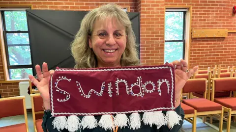 Semra Heath smiles at the camera while holding a fabric banner made for the project with Swindon embroidered on it