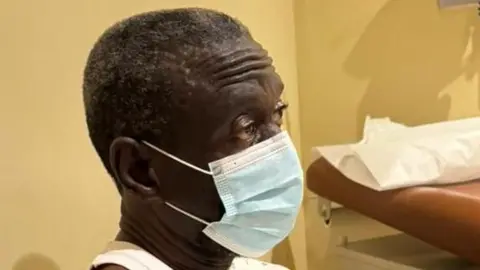 Kizza Besigye seated on a chair in a clinic room. He is wearing a mask and a hospital gown in a Kampala medical facility 