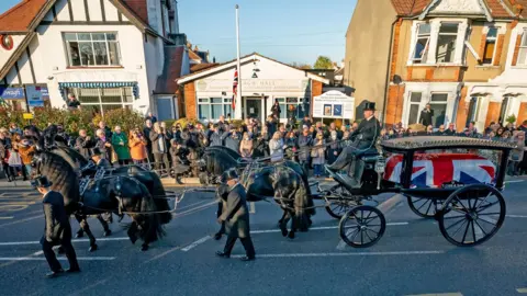 Aaron Chown/PA A horse-drawn funeral cortege passes along a residential street. Sir David Amess's coffin is draped in a union jack in glass-sided carriage. People line the pavement beyond the procession. The local Conservative Party office is behind them, with houses on either side.