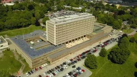 An aerial picture of County Hall in Norwich, the headquarters of Norfolk County Council. It is a large multi-floored building with cars in front and set amid trees and a grassed area.