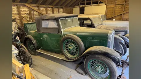 Humbert & Ellis A 1932 Alvis 1250 sitting in a barn. It has a dark green body with a black soft top roof, with black tyres and a spare black tyre against the bonnet, plus a green jump seat at the back. It is covered in dust and behind it are two other dust-covered vintage cars.