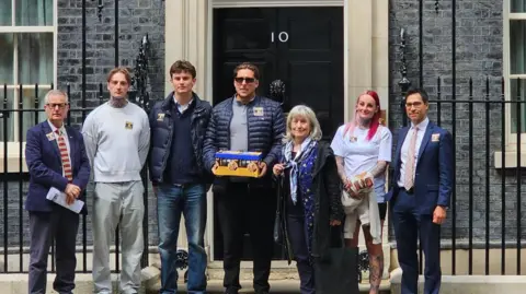 Un grupo de personas se encontraba frente a la puerta negra del número 10 de Downing Street. Joe Bennett, en el centro, lleva una caja amarilla y azul.